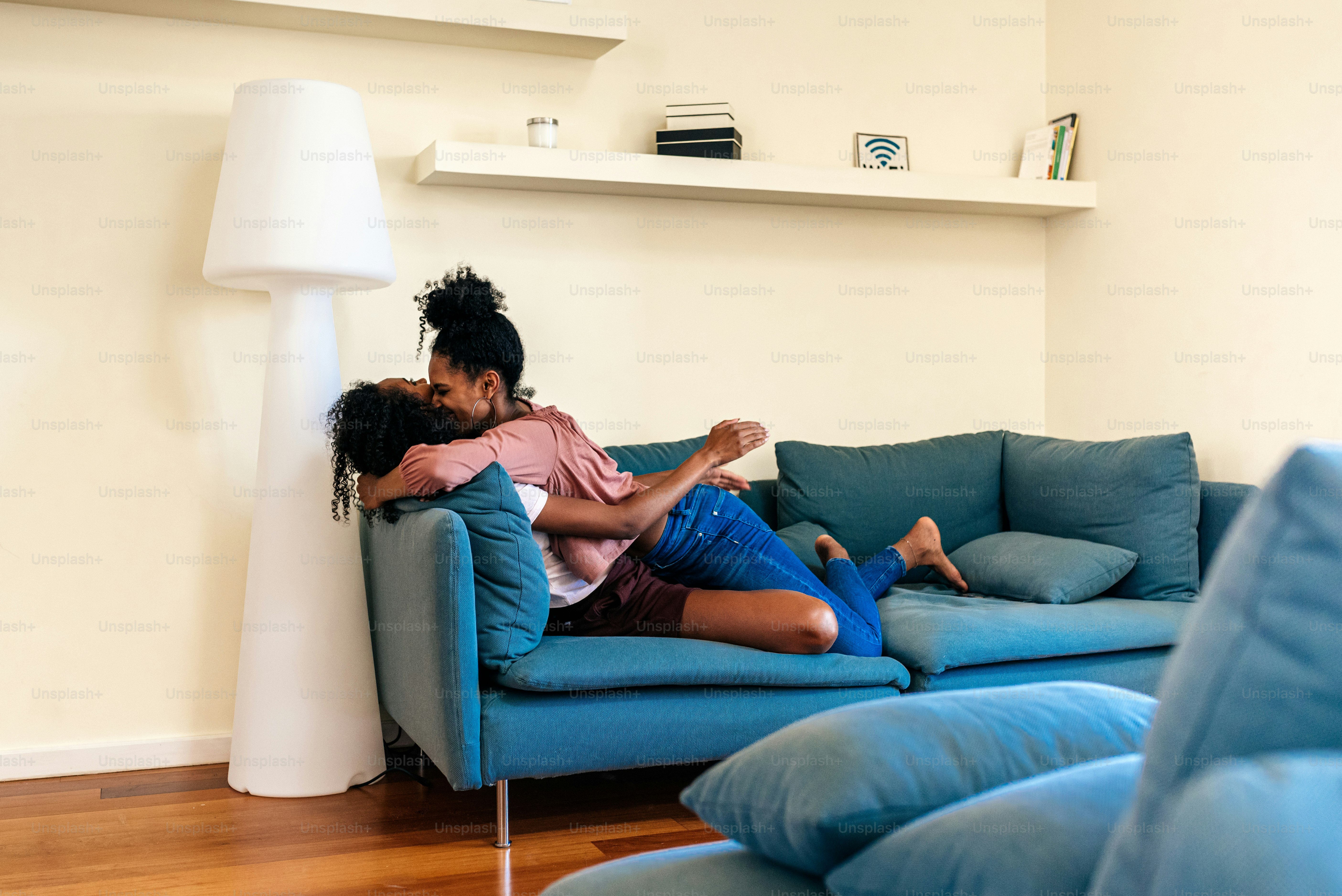 Overjoyed African American females in casual clothing laughing while smiling and cuddling on blue cozy sofa at home
