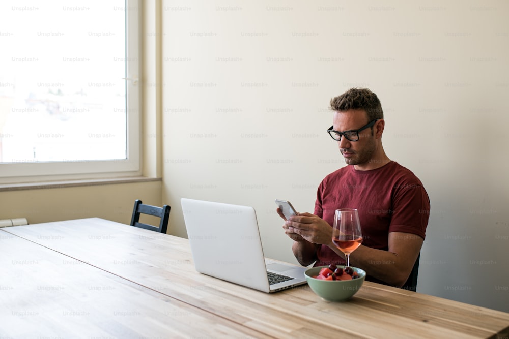 Focused blonde man browsing laptop and smartphone at wooden table with glass of wine and bowl with berries at home