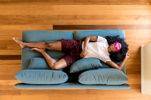 Top view of African American woman in home clothing having relaxation on blue couch with pink eye mask at home