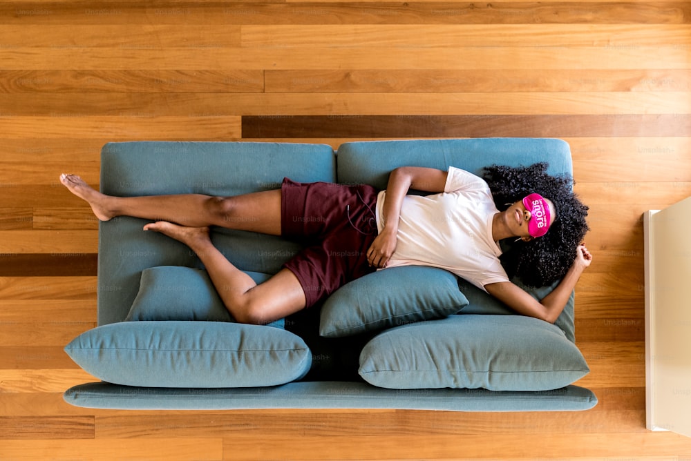 Top view of African American woman in home clothing having relaxation on blue couch with pink eye mask at home