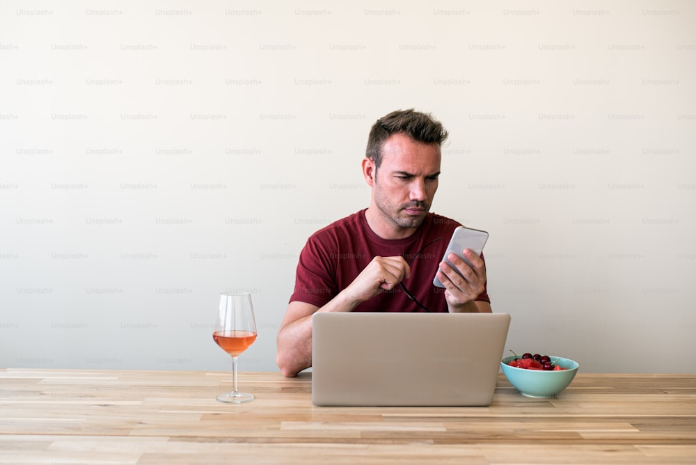 Fokussierter blonder Mann durchstöbert Laptop und Smartphone am Holztisch mit Glas Wein und Schüssel mit Beeren zu Hause