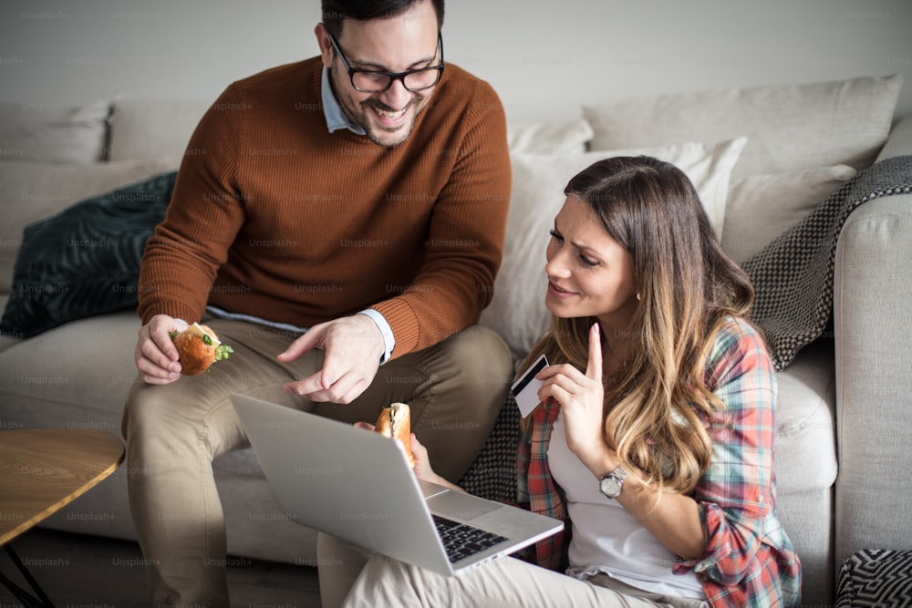 Checking the account balance. Happy couple at home using laptop and eating sandwich.