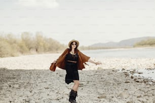 happy stylish hipster traveler woman in hat, fringe poncho walking near water river beach in mountains, gypsy boho girl. wanderlust summer travel. atmospheric moment. space for text.