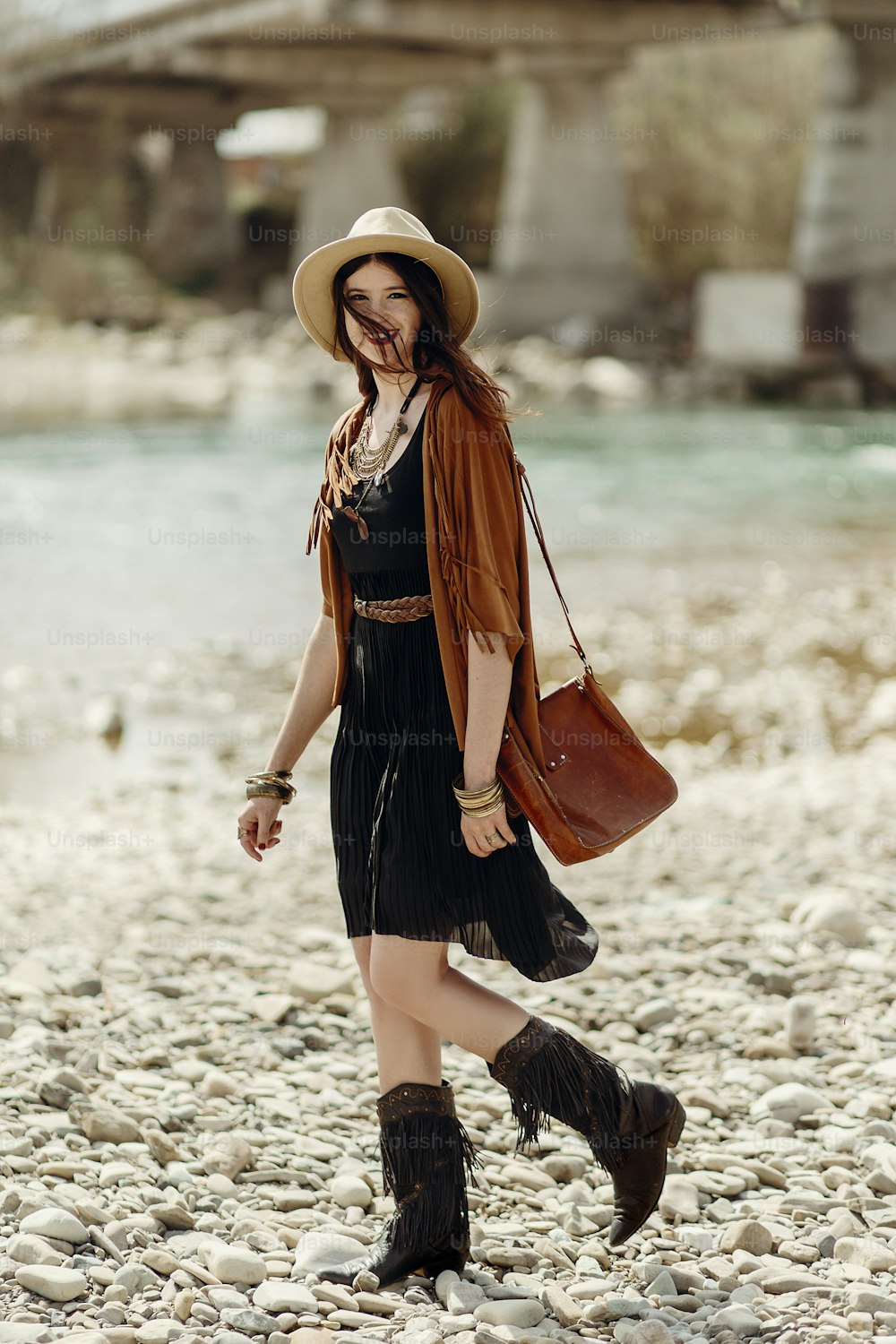 stylish hipster woman in hat, fringe poncho and boots walking on river beach. boho traveler girl in gypsy look, summer travel. atmospheric moment.  space for text.