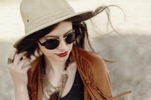 stylish boho woman in sunglasses holding hat, with windy hair. hipster girl in gypsy look, young traveler near river beach in mountains. atmospheric moment. summer travel. space for text.