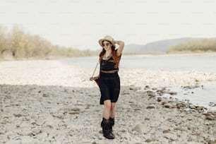 stylish boho traveler woman in hat, fringe poncho walking near water river beach in mountains, gypsy hipster girl. wanderlust summer travel. atmospheric moment. space for text.