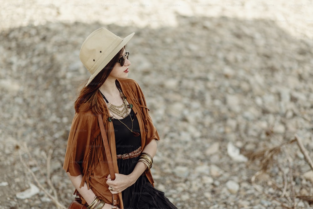 beautiful stylish boho woman in sunglasses and hat, fringe poncho and leather hat. hipster girl in gypsy look young traveler posing near river beach in mountains.  atmospheric moment