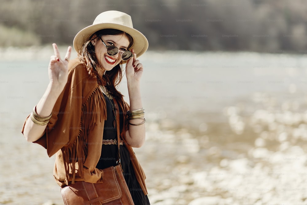 stylish hipster boho woman smiling showing peace sign in sunglasses with hat, leather bag, fringe poncho and accessory. happy traveler girl look, wanderlust summer travel. space for text