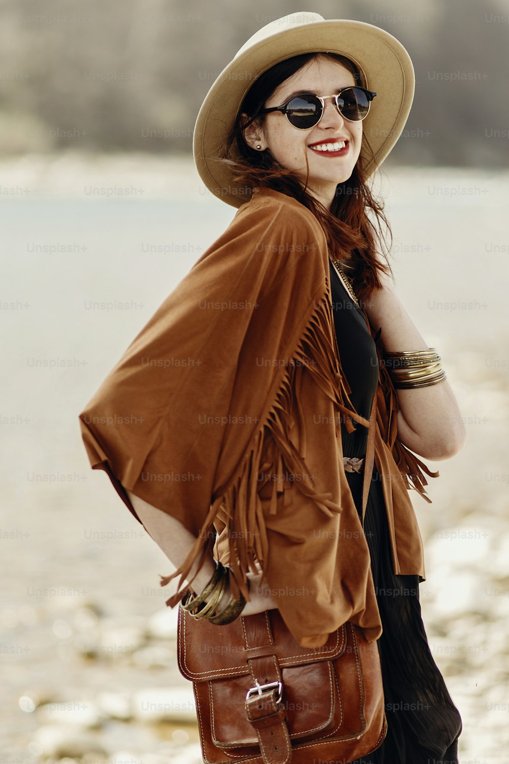 stylish hipster boho woman smiling in sunglasses with hat, leather bag, fringe poncho and accessory. happy traveler girl look, near beach in mountains. wanderlust summer travel.