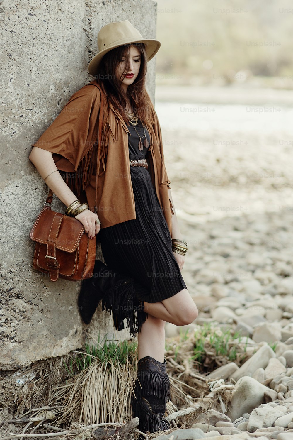 beautiful stylish boho woman with hat, leather bag, fringe poncho and boots. girl in gypsy hippie look young traveler posing near river rocks in mountains. sensual look. atmospheric moment