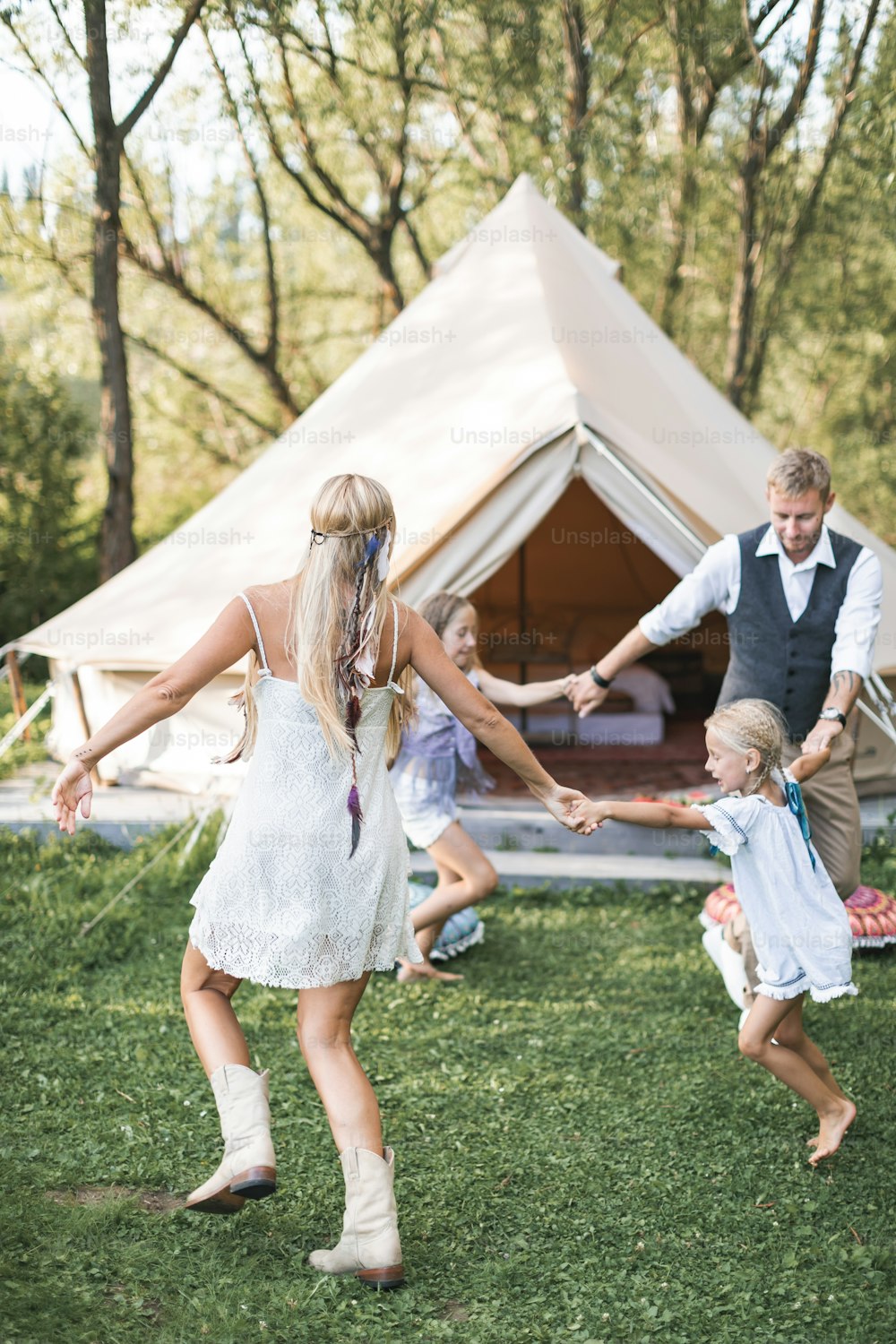 Happy active family wearing stylish boho casual clothes, dancing and jumping in green field or park in front of tipi tent. Summer vacation, family concept.