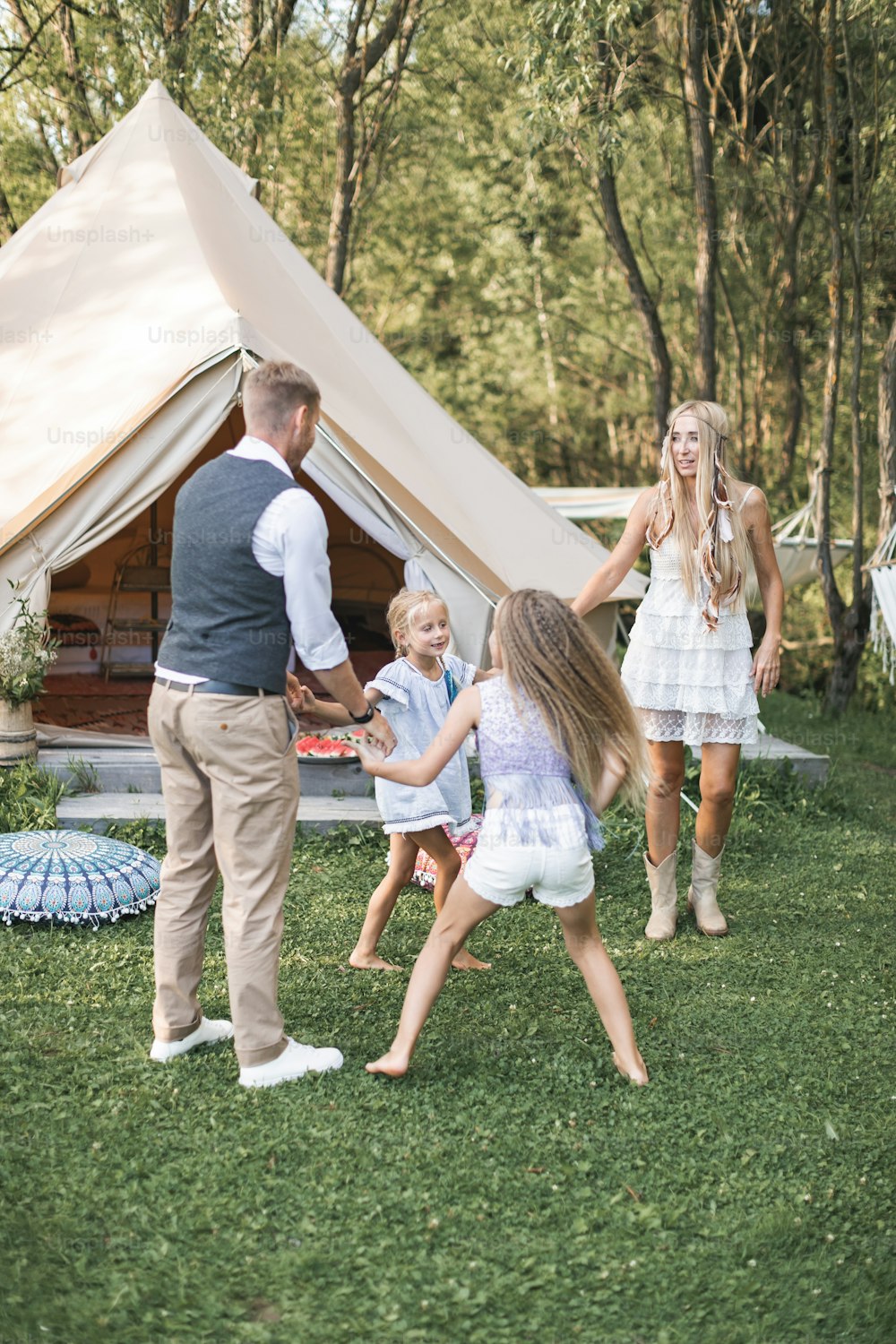 Young family is playing together on a sunny day in park. Father, mother and two daughters in casual boho clothes dancing and jumping, holding hands in front of big white tipi wigwam tent.