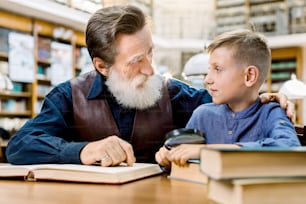Petit garçon souriant heureux avec son grand-père barbu joyeux lisant des livres à la bibliothèque, se regardant. Petit garçon souriant avec son professeur principal étudiant ensemble dans une bibliothèque vintage.