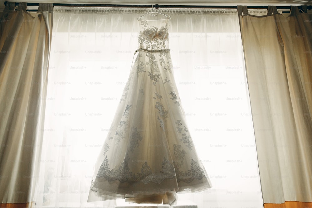 Schönes weißes Brautkleid in der Nähe eines Fensters im Hotelzimmer, morgendliche Hochzeitsvorbereitung, weißes Spitzenkleid für Braut auf Kleiderbügel