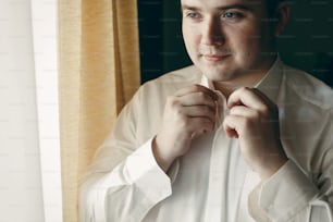 Handsome young man preparing for business meeting, putting on shirt, happy groom posing near window in hotel room before the wedding, businessman concept
