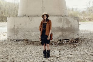 stylish hipster woman in hat and fringe poncho and boots posing near river stones. boho traveler girl in gypsy look. summer travel. atmospheric moment. space for text.