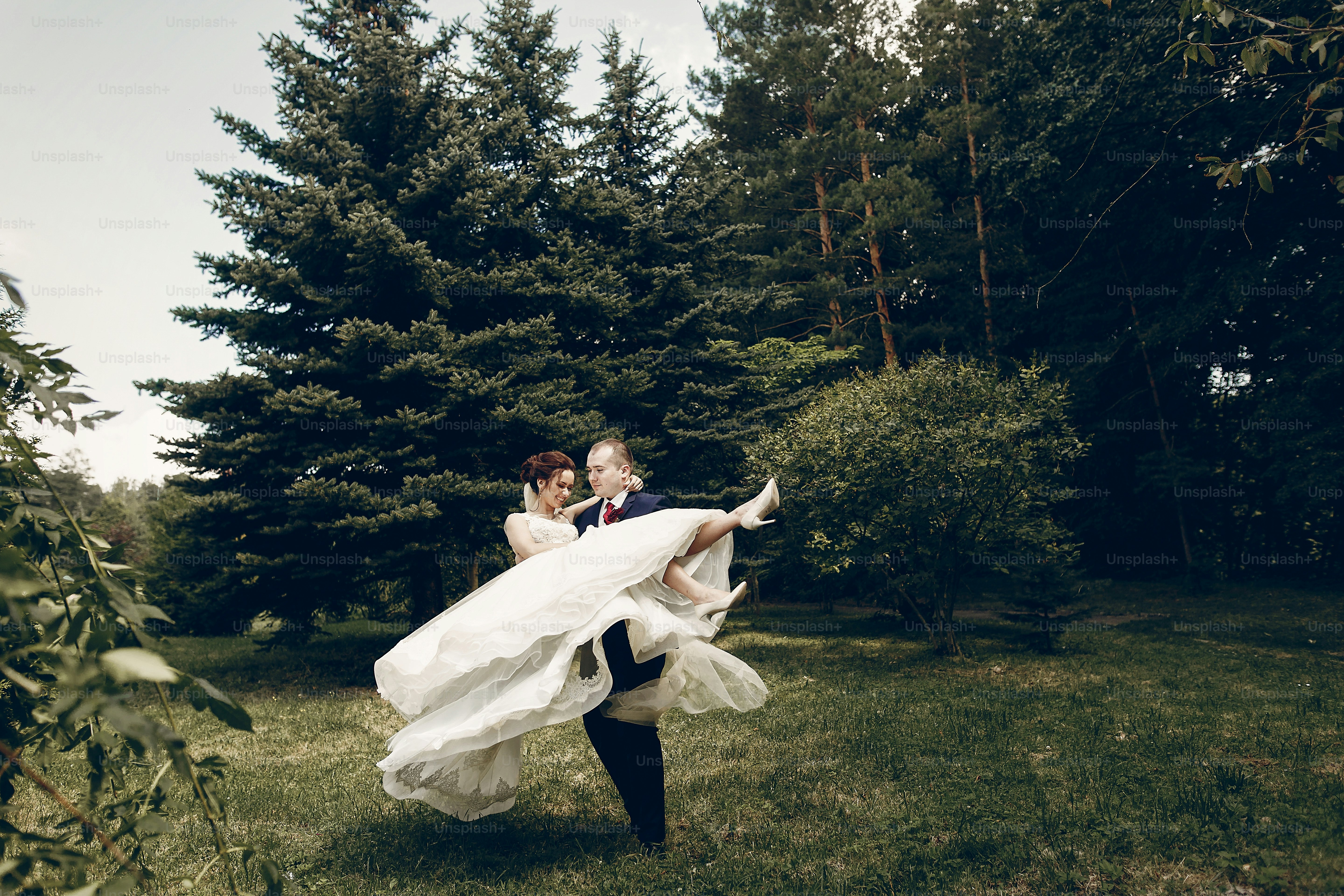 Handsome strong groom holding happy bride in the air, newlywed couple posing in summer park, groom carrying beautiful bride, romantic moment concept