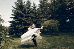 ハンサムな強い花婿が幸せな花嫁を空中に抱えている、新婚カップルが夏の公園でポーズをとっている、花婿が美しい花嫁を運ぶ、ロマンチックな瞬間のコンセプト