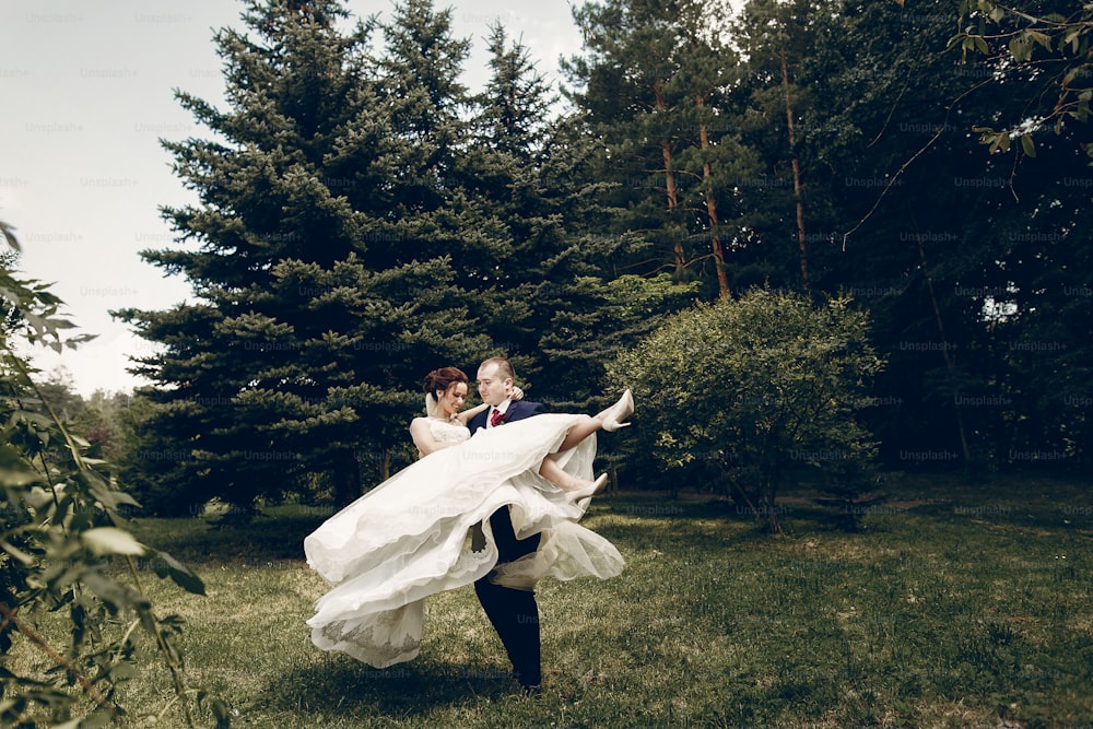 Handsome strong groom holding happy bride in the air, newlywed couple posing in summer park, groom carrying beautiful bride, romantic moment concept