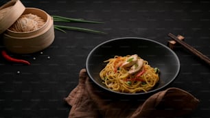 Cropped shot  of Schezwan Noodles or Chow Mein with vegetable and chicken served in black bowl and ingredients on black table