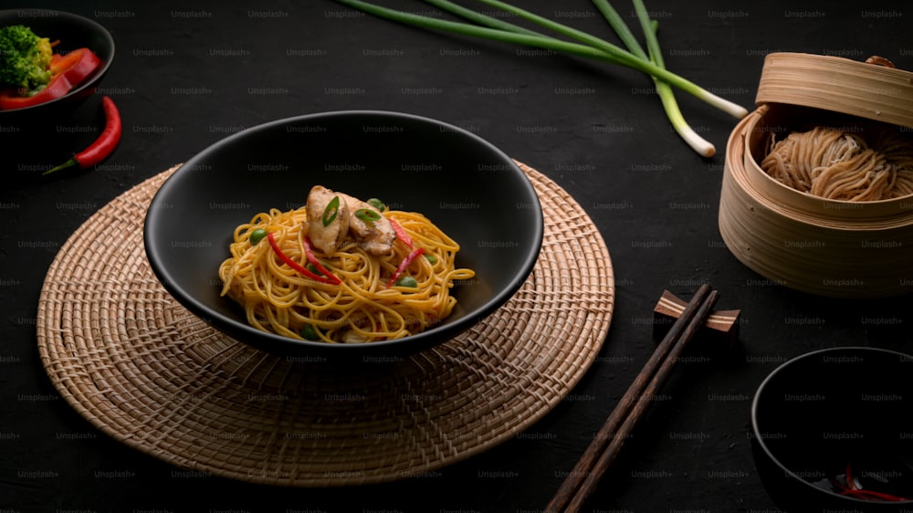 Cropped shot  of Schezwan Noodles or Chow Mein with vegetable, chicken and chilli sauce served in black bowl and ingredients on black table