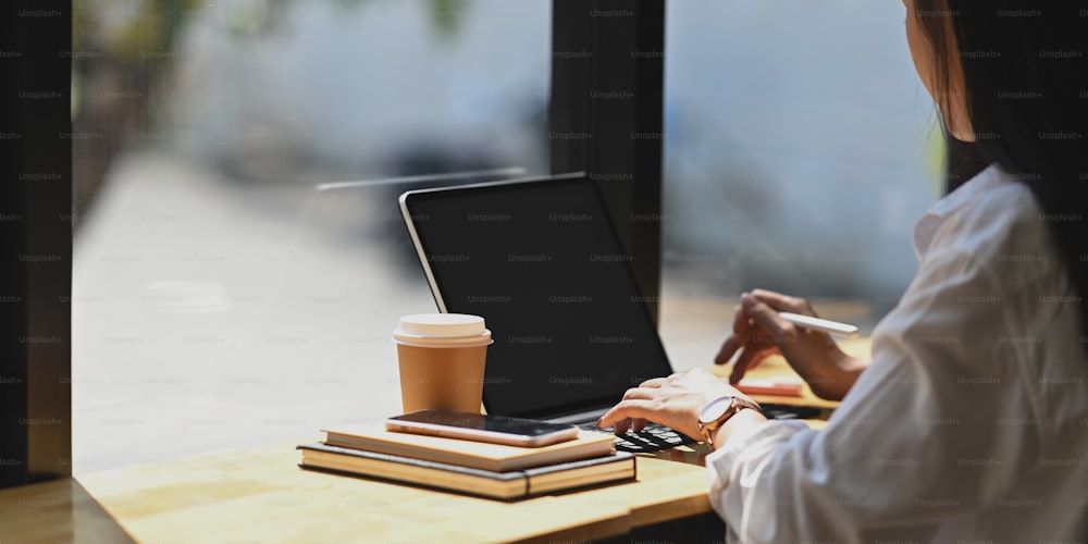 흰색 셔츠를 입은 아름다운 사업가가 키보드 케이스가 있는 컴퓨터 태블릿에 타이핑하는 동안 카페/레스토랑 창문이 있는 나무 작업 테이블에 앉아 있는 측면 촬영 이미지.