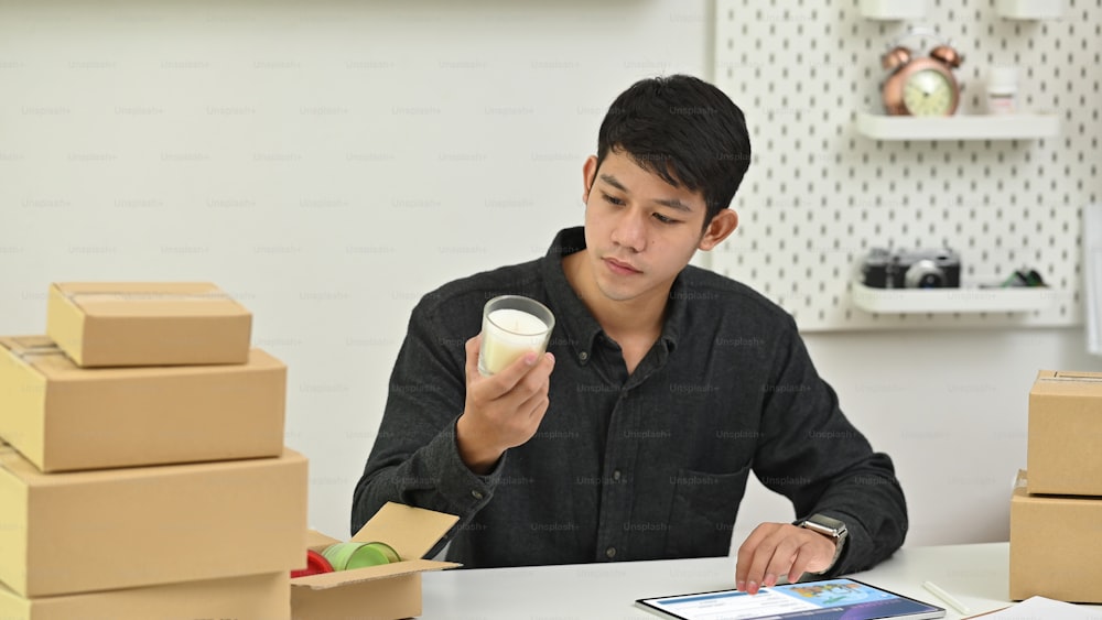 Porträt eines jungen Geschäftsinhabers, der seine Waren packt, während er am modernen Schreibtisch sitzt, umgeben von einem Stapel von Pappkartons mit gemütlichem Wohnzimmer als Hintergrund.