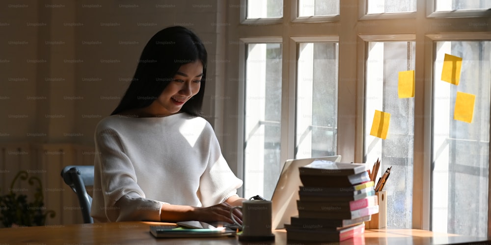 整然とした図書館のフロントコンピュータラップトップ、コーヒーカップ、本やノートのスタックの木製のテーブルに座って、彼女の手にスマートフォンを保持している美しい女性の写真。