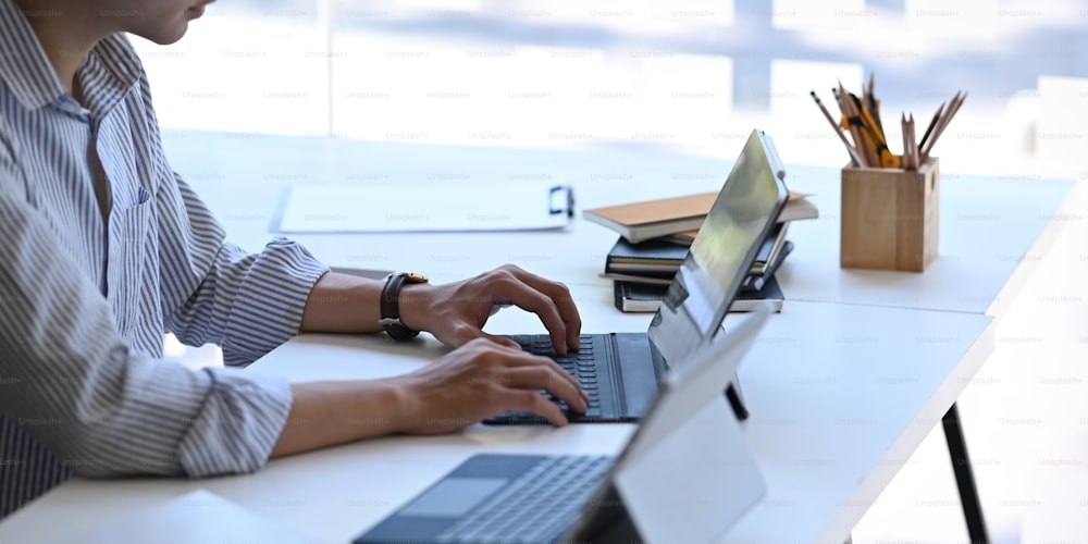 Photo of young businessman typing on computer tablet with keyboard case while sitting in front two of computer tablet, stack of books, pencil holder and clipboard that all of them putting on table.