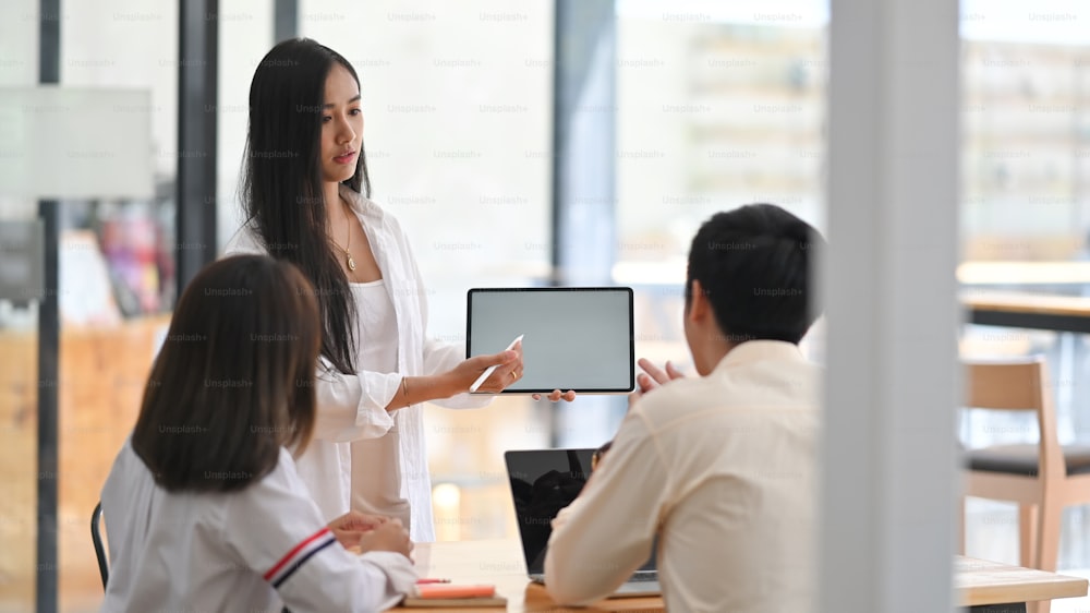 Side shot of young beautiful woman in white shirt showing a computer tablet and stylus pen while standing at the meeting table with her colleague in modern office as background.