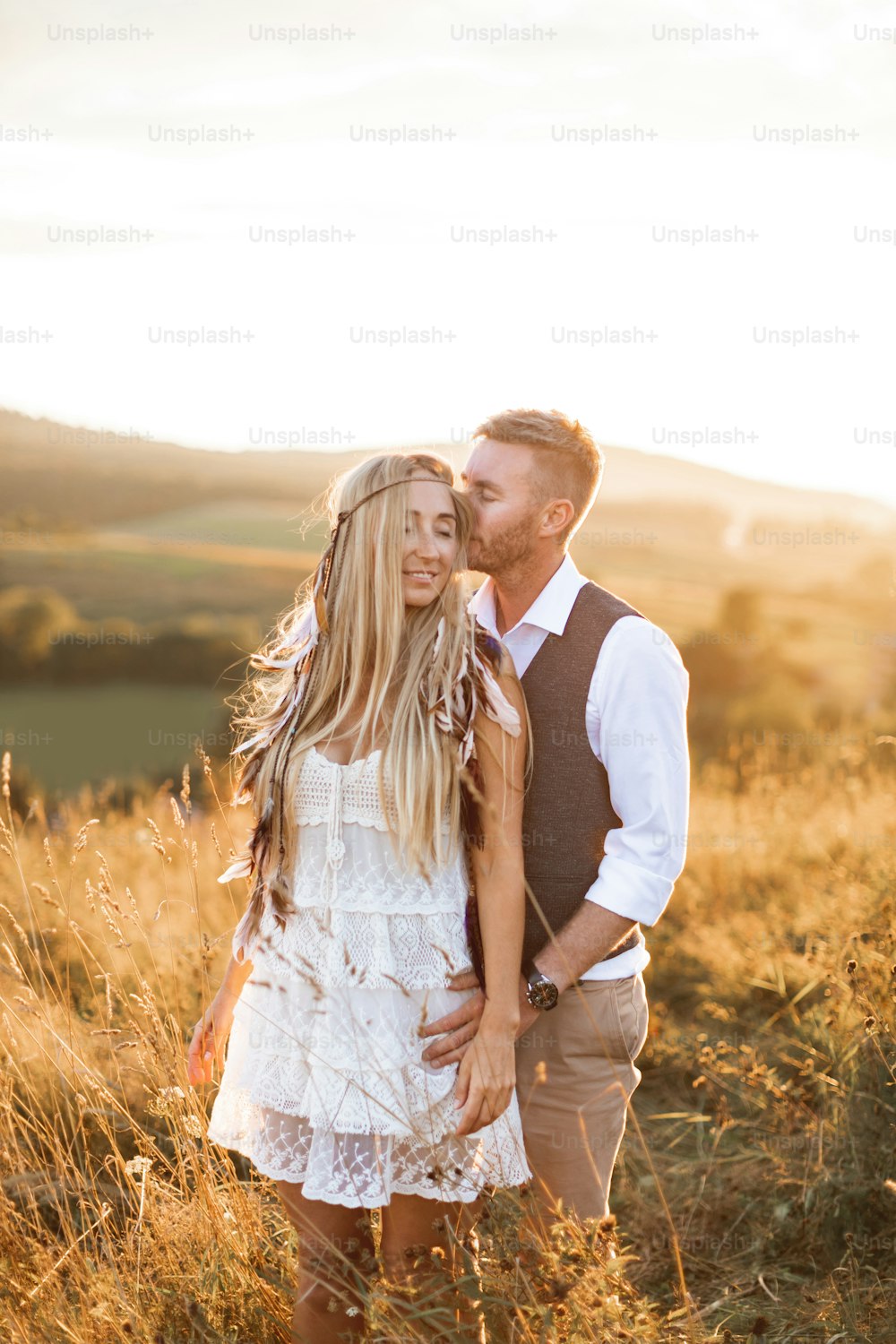 Pareja boho tomada de la mano y abrazándose juntos, caminando en el campo rural. Mujer bonita con vestido blanco y accesorios de plumas boho en el pelo, hombre guapo con traje casual.