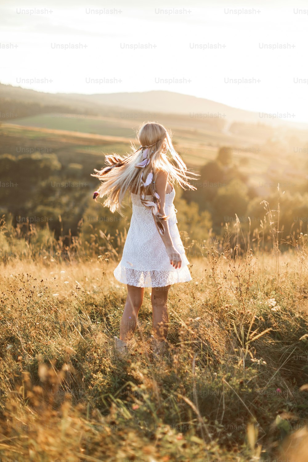 Young hipster girl in white dress and hair feathers standing outdoors, in summer field in mountains. Boho hippie style.