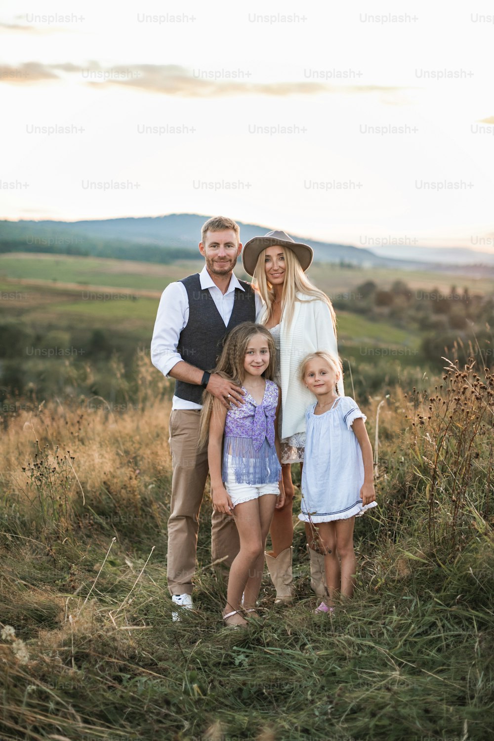 Full length portrait of cheerful Caucasian family, mother, father and two little daughters, standing in summer wild field, mountains on the background.