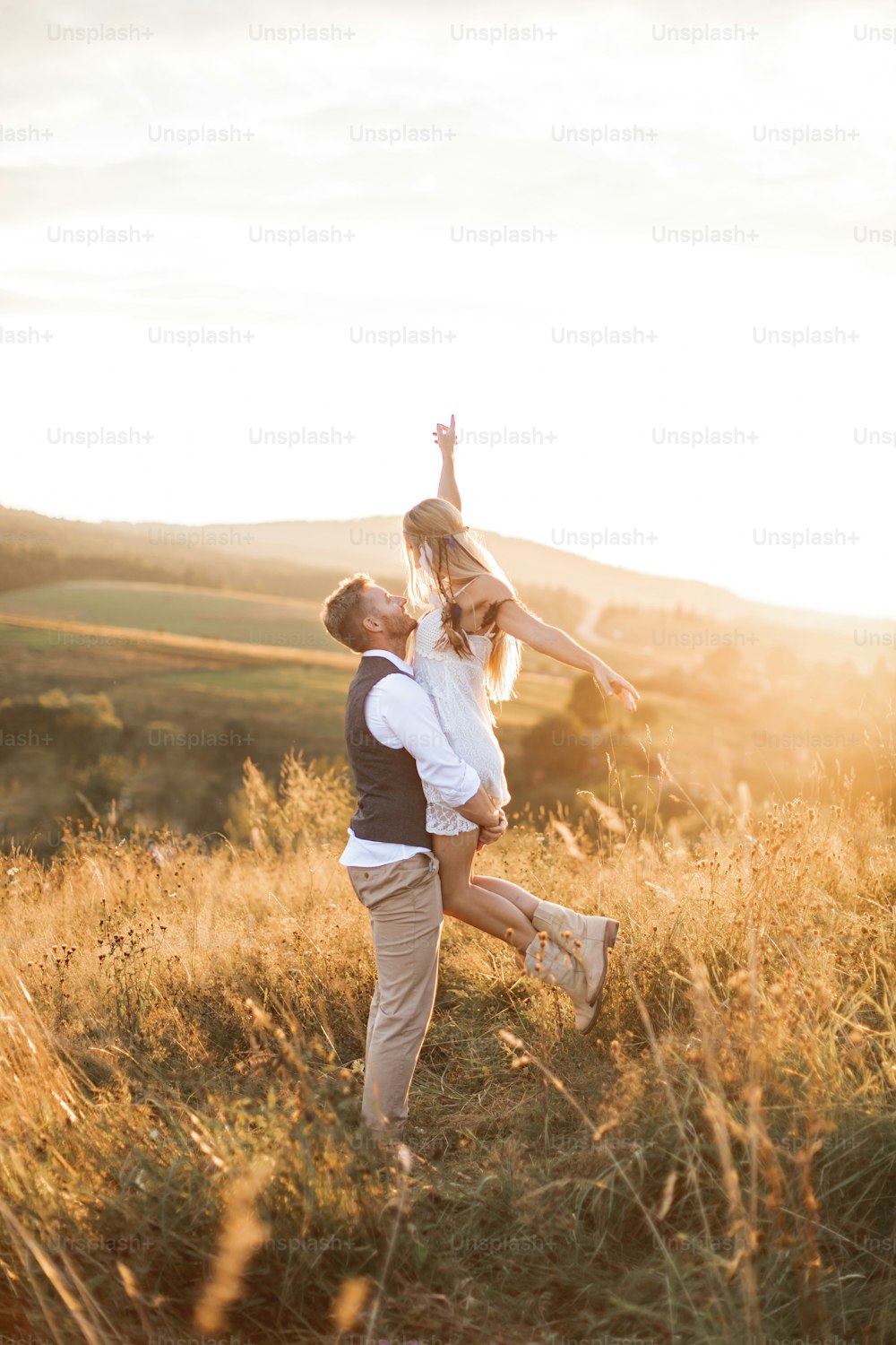 Shot of young boho hippie woman being carried by her handsome boyfriend in summer field. Couple having fun on their summer evening outdoors.