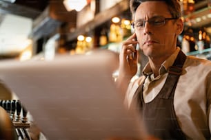 Mid adult barista reading paperwork while communicating with someone over mobile phone in a bar.