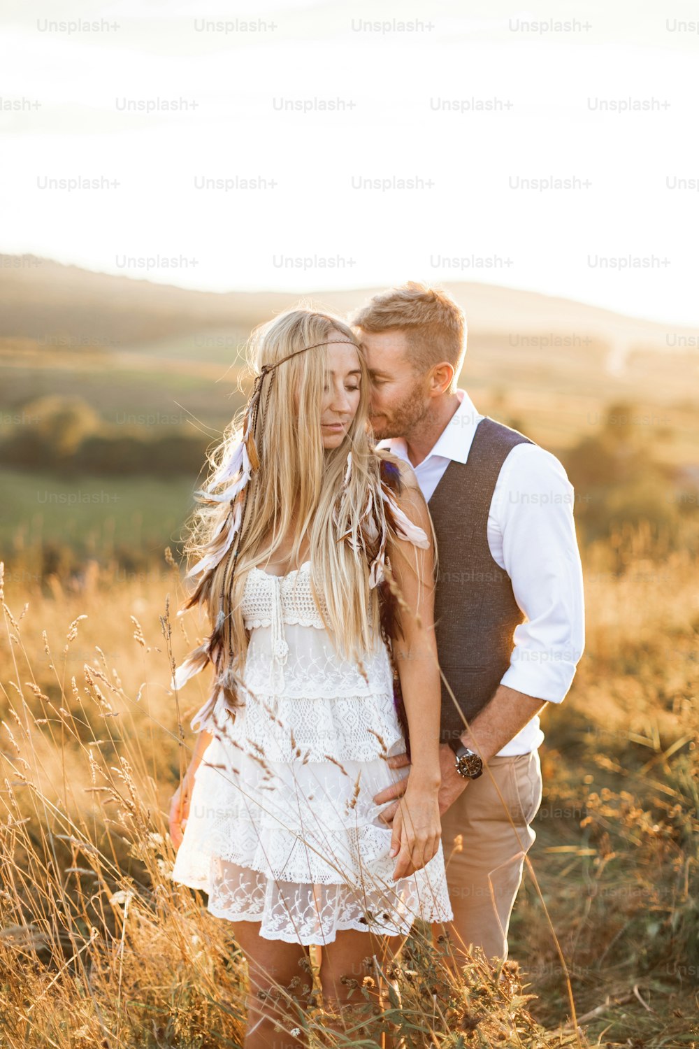 boho couple holding hands and embracing together, walking in countryside field. Pretty woman in white dress and boho feather accessories in hair, handsome man in casual suit.