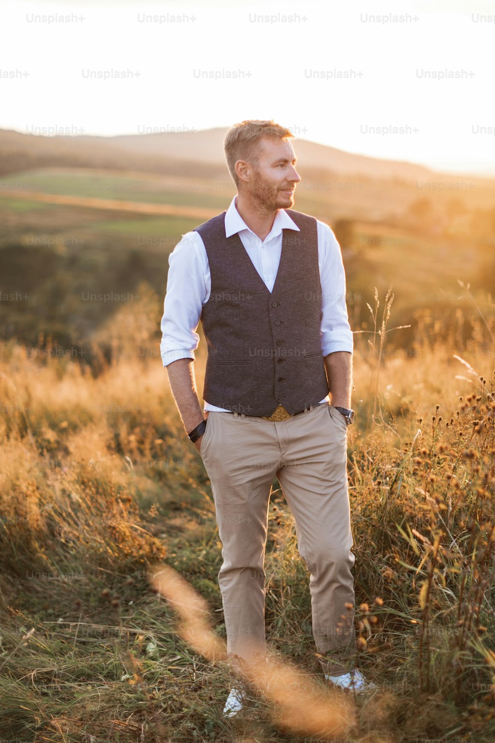 Portrait of handsome young man, farmer or cowboy, wearing casual suit, looking to a side, while standing in summer field in countryside, sunset.
