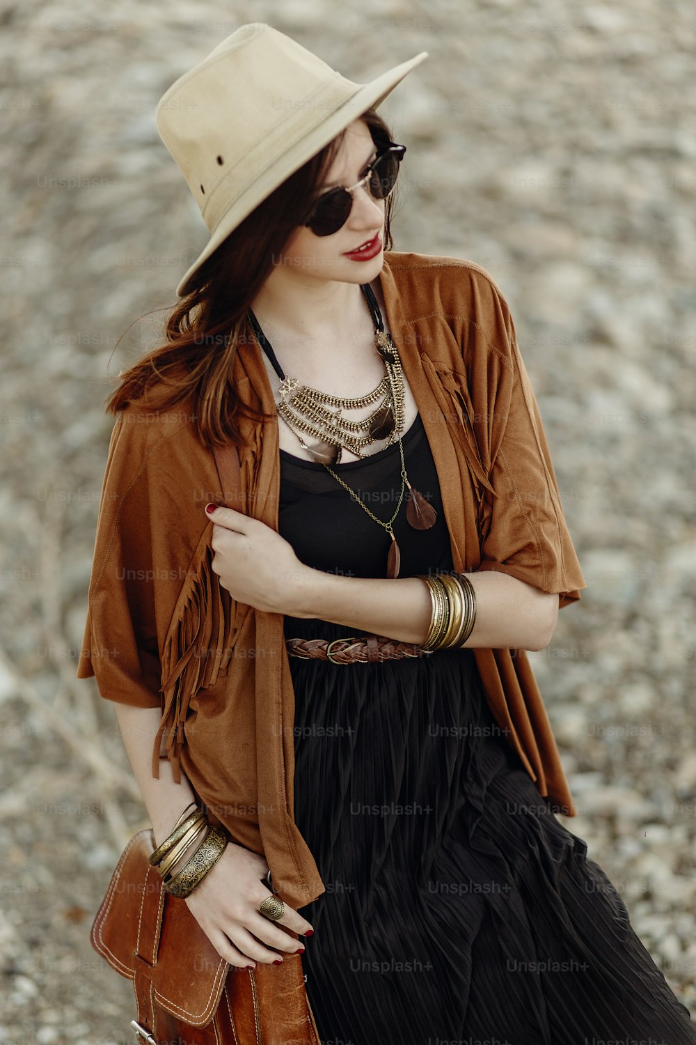 beautiful stylish boho woman in sunglasses and hat, fringe poncho and leather bag. hipster girl in gypsy look young traveler posing near river beach in mountains. sensual look.