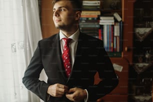 Hübscher Bräutigam im weißen Hemd mit roter Krawatte zugeknöpfter schwarzer Anzug, morgendliche Hochzeitsvorbereitung, Geschäftsmann im Anzug schaut aus dem Fenster
