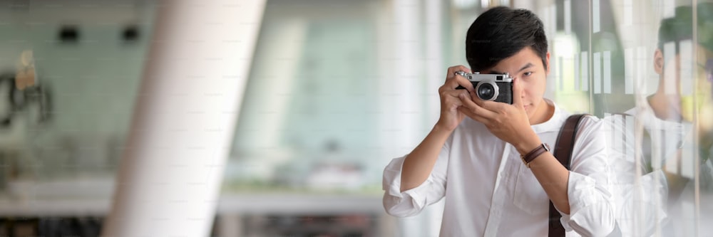 Beschnittene Aufnahme eines jungen männlichen Fotografen, der mit der Digitalkamera fotografiert, während er in einem Büro mit Glaswand steht