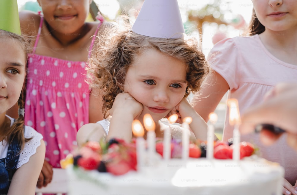 Um retrato de crianças com bolo em pé ao redor da mesa na festa de aniversário no jardim no verão.