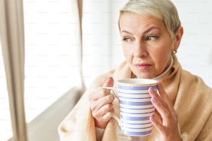 Waist-up portrait of a dreamy attractive aged woman with a mug of tea looking away