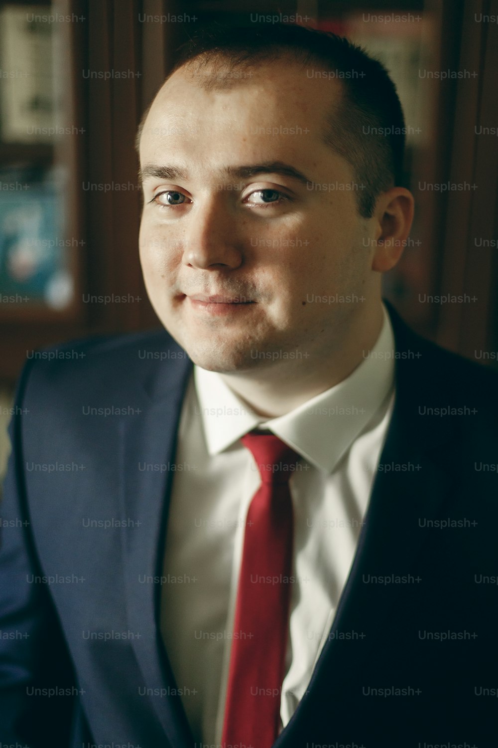 Glücklicher stilvoller Bräutigam im dunkelblauen Anzug mit roter Krawatte posiert morgens vor der Hochzeit im Hotelzimmer, Porträt eines glücklichen Geschäftsmannes drinnen