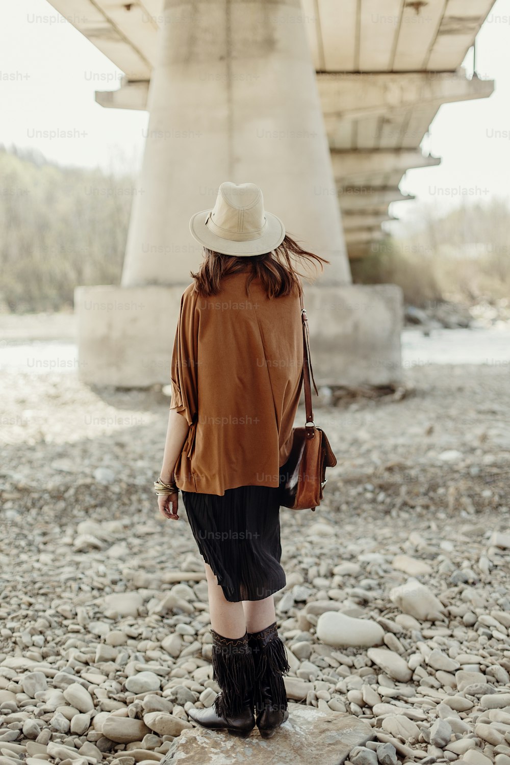 stylish boho woman posing at river, back view. beautiful gypsy girl with hat and fringe poncho and boots. young girl traveler. fashionable hippie outfit. atmospheric moment. space for text