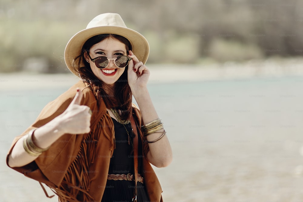 Femme hipster voyageuse élégante souriante montrant le pouce levé, en lunettes de soleil avec chapeau, sac en cuir, poncho à franges et accessoire. Look de fille bohème heureuse, voyage d’été Wanderlust. espace pour le texte.