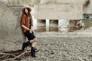 stylish boho woman with jewelry posing at rock wall. beautiful gypsy dressed girl with hat and fringe poncho with sensual look. young girl traveler. fashionable hippie outfit. space for text