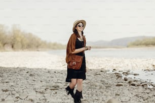happy stylish hipster traveler woman in hat, fringe poncho walking near water river beach in mountains, gypsy boho girl. wanderlust summer travel. atmospheric moment. space for text.