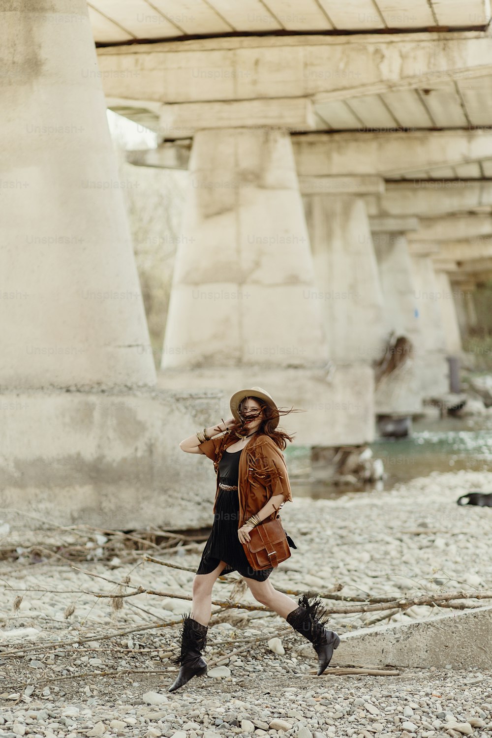 stylish boho woman jumping, having fun, in hat, leather bag, fringe poncho and boots  near river under bridge stone. girl in gypsy hippie look young traveler. summer travel