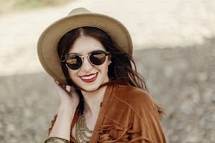 stylish boho woman smiling in sunglasses holding hat, with windy hair. hipster girl in gypsy look, young traveler near river beach in mountains. atmospheric moment.  space for text.