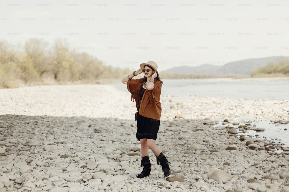 happy stylish hipster traveler woman in hat, fringe poncho walking near water river beach in mountains, gypsy boho girl. wanderlust summer travel. atmospheric moment. space for text.